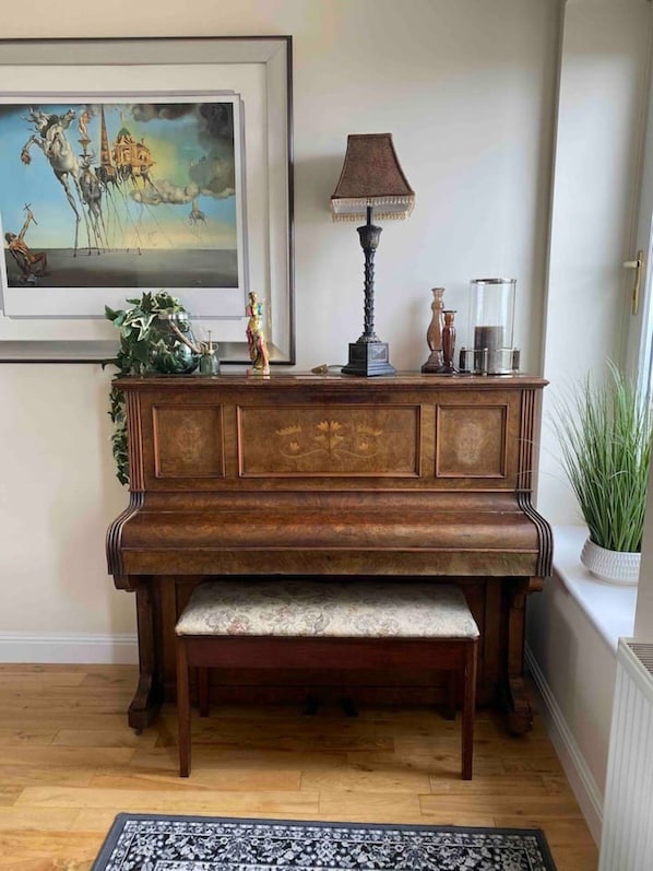 Un piano de cola con un banco en una habitación con piso de madera, un cuadro en la pared, una lámpara y objetos decorativos sobre una consola.