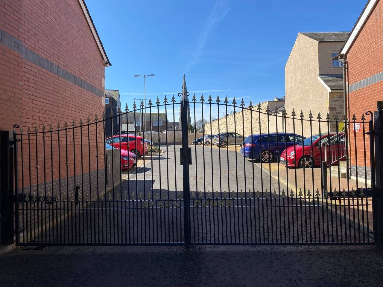 A gated parking area with multiple cars parked, a clear sky, and a building with a brick facade in the background.