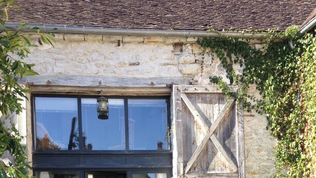 Un bâtiment en pierre doté d’une grande porte et de grandes fenêtres en verre, entouré d’un jardin avec un sentier en pierre et diverses plantes.
