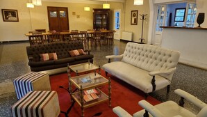 A lobby with a dark brown sofa, a white armchair, striped ottoman, and a glass coffee table with magazines. There are wooden chairs and tables in the background, and a coat rack is visible. - Bosque De Bohemia Delta Tigre (Partido de Tigre)