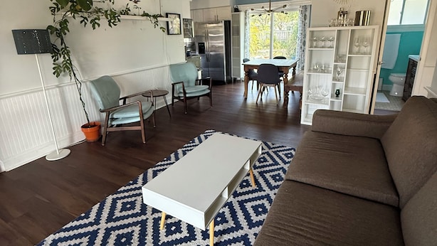 Un soggiorno moderno con un divano marrone, un tavolino da caffè bianco e un tappeto azzurro con motivi. Si intravede una ventola a soffitto, un'area pranzo con sedie e una cucina sullo sfondo.