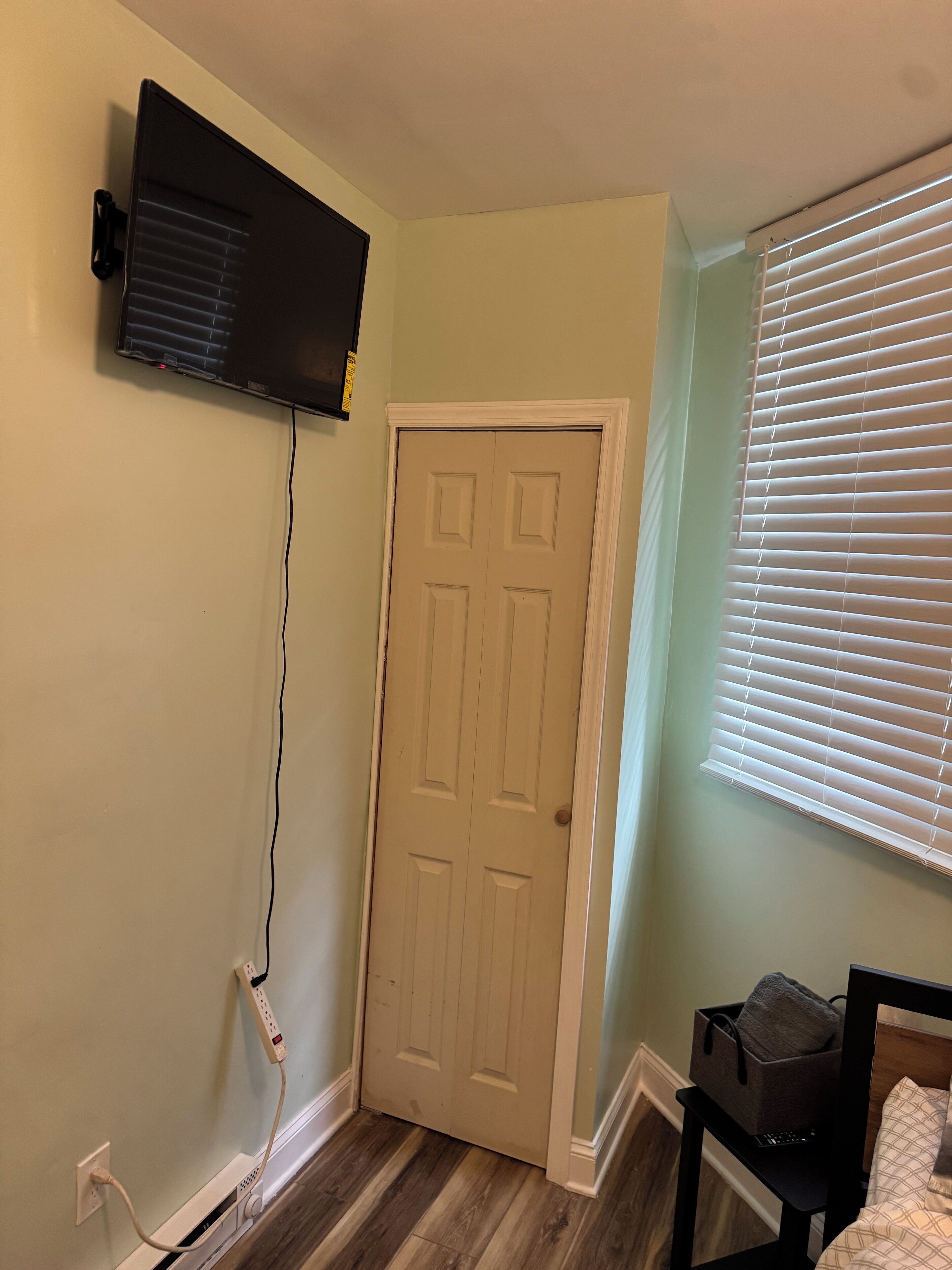 A room with a closed door, a mounted television, a power outlet, a window with blinds, and a small table with a bag.