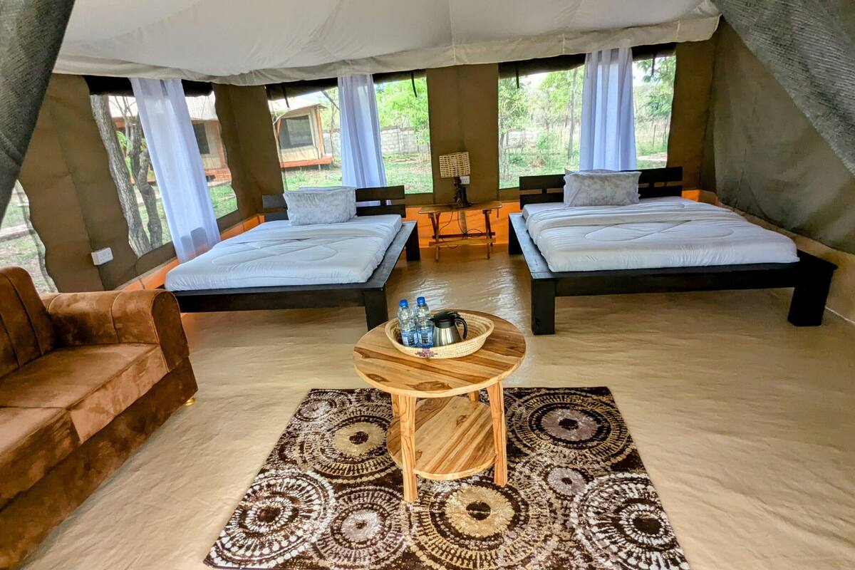 Un hébergement de type tente comprenant deux lits, un canapé, une table avec des bouteilles d’eau et un tapis à motifs.