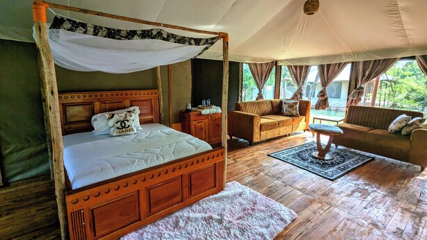 A four-poster bed with a canopy, a sofa, a small table, and a rug in a room with wooden floors and large windows. - Double G Safaris And Camp Mikumi (Dar es Salaam)