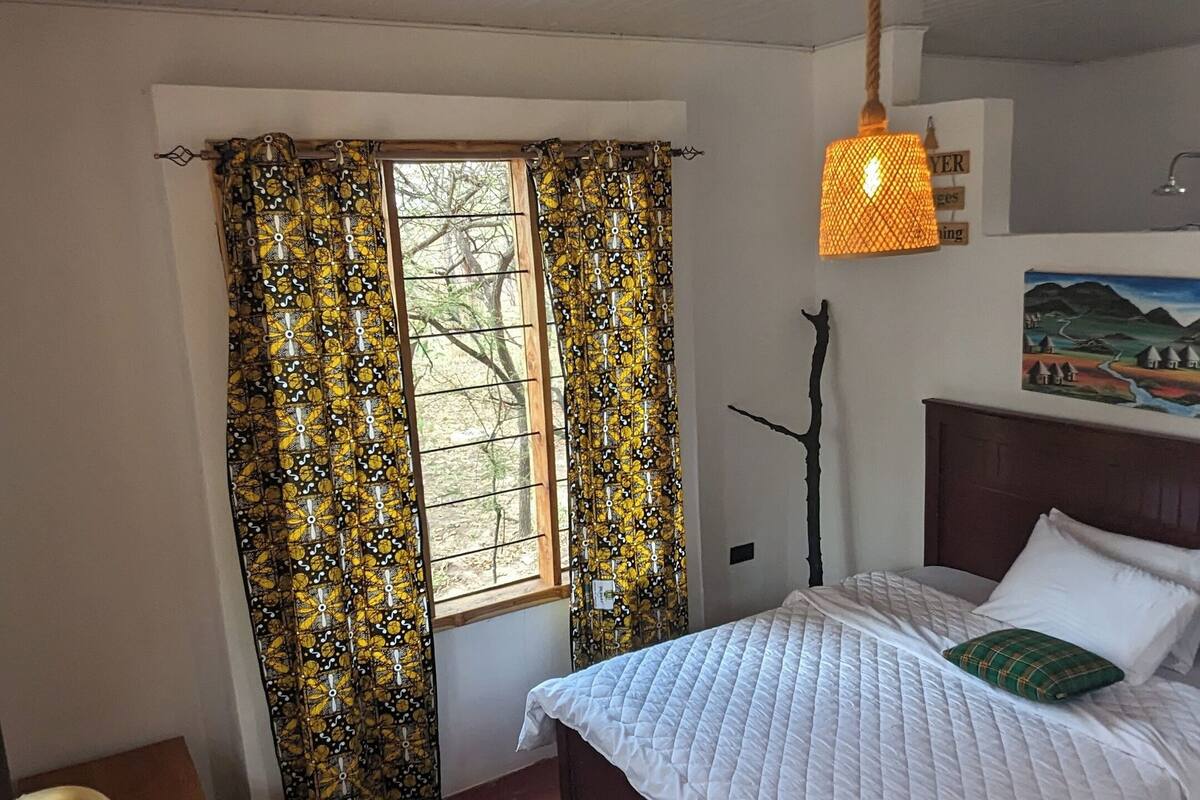 Une chambre à coucher avec un lit, une chaise, une fenêtre avec des rideaux, un tableau au mur et un luminaire suspendu.