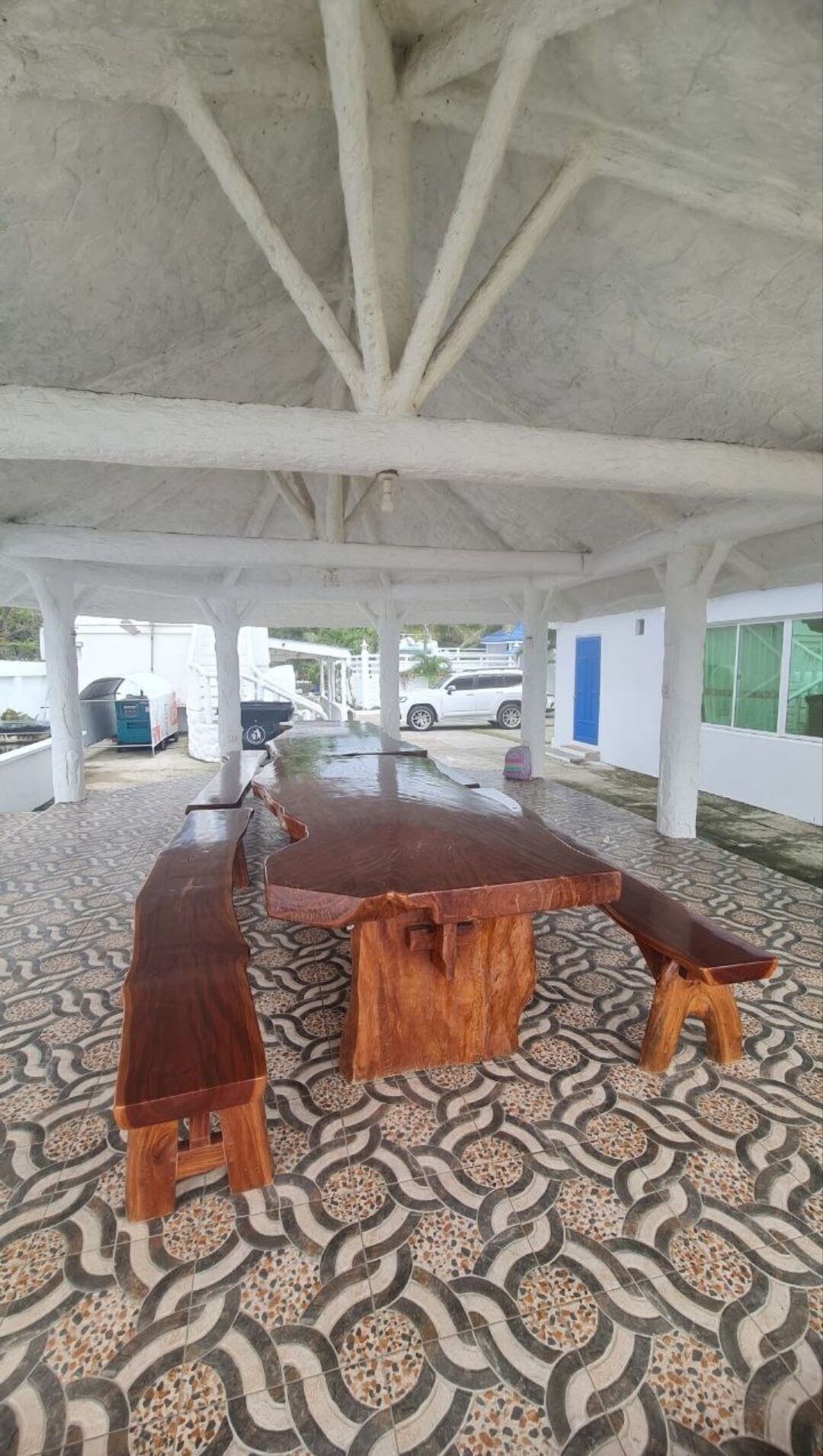An outdoor covered area with a long wooden table and benches, surrounded by patterned flooring and white pillars.