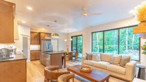 A modern kitchen with wooden cabinets, a central island, and pendant lighting. There is a dining area with a wooden table and chairs, and a living room with a sofa and large windows offering a view of the outdoors. - Luxury Mountain-Modern Retreat 3BR near Glacier National Park & Whitefish (Columbia Falls)