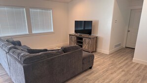 Un salon avec un canapé d’angle gris, un meuble de rangement en bois abritant un téléviseur, et une porte donnant sur une autre pièce.