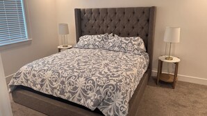 Une chambre à coucher avec une tête de lit capitonnée, une couvre-lit à motifs, deux tables de chevet avec des lampes et une fenêtre avec des stores.