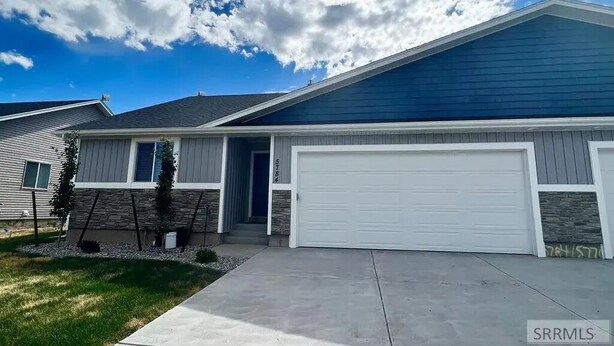 Une maison de plain-pied avec un garage, des fondations en pierre et un ciel bleu dégag é parsemé de nuages.