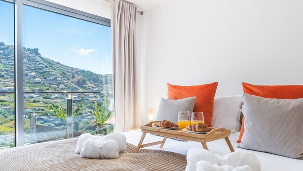 A hotel room with a large bed, a breakfast tray with croissants and orange juice, and a view of a hilly landscape.
