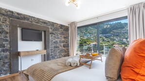 A modern hotel room with a stone wall, a flat-screen TV, a wooden bedside table with a tray of breakfast, and a large window with a view of a hillside town.