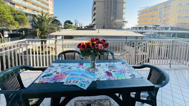 A table with a floral tablecloth and a vase of flowers, set on a balcony with a view of a hotel building. - Comfortable Flat a few Steps From the Beach (San Michele al Tagliamento)