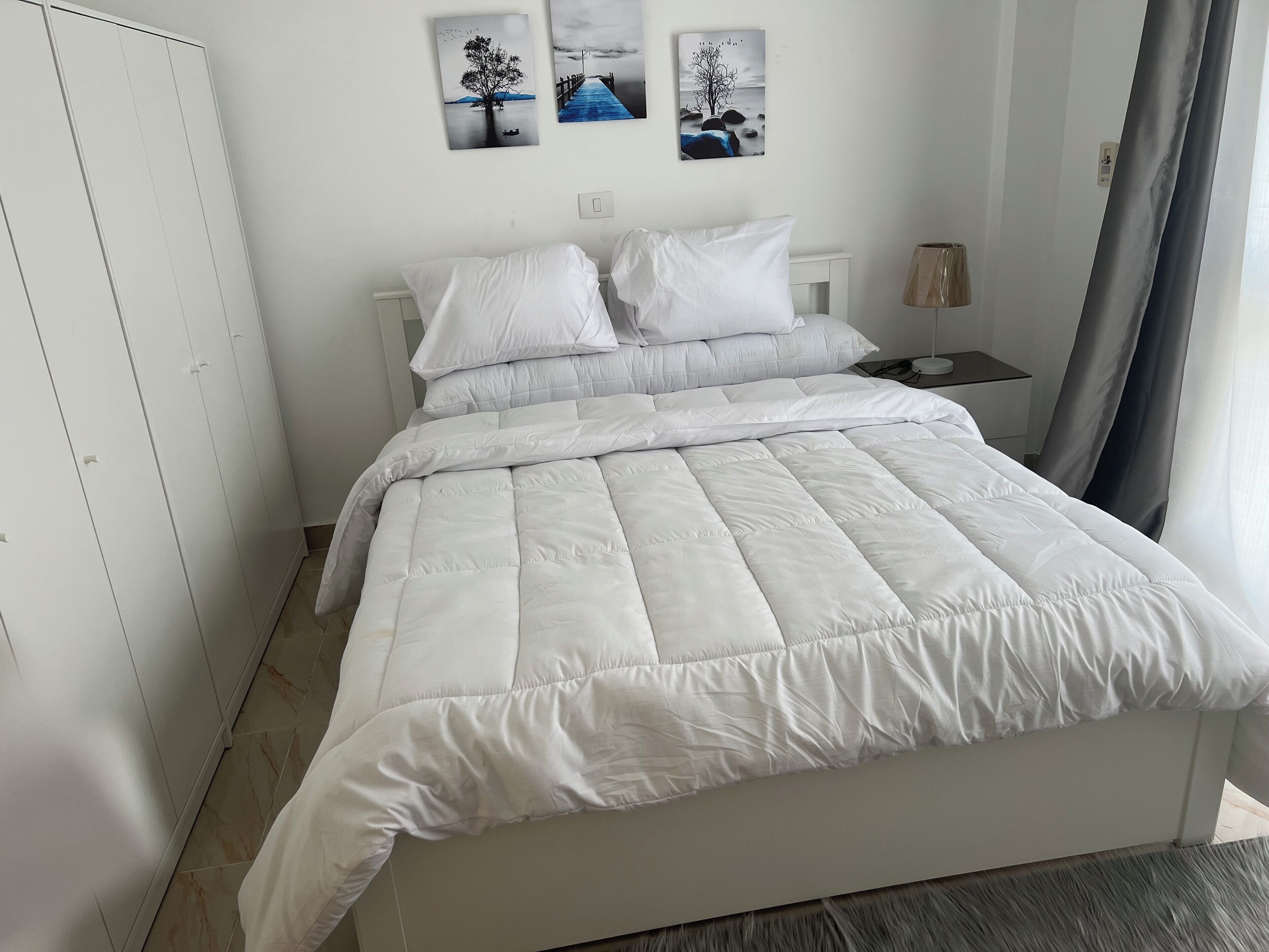 Een opgemaakt bed met witte beddengoed, een witte kledingkast en drie kunstwerken aan de muur met afbeeldingen van bomen en landschappen.