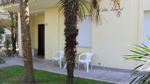 A building with a yellow facade, a palm tree in the foreground, and white plastic chairs outside. - Luminous Flat Near the Beach (San Michele al Tagliamento)