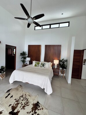 A bedroom with a bed, ceiling fan, wooden headboard, bedside tables, lamps, and a cowhide rug. - Villa Montemar - Mountain and Ocean Views + Infinity Pool (Platanillo)