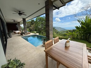 A poolside area with a wooden table and chairs, a ceiling fan, and a view of the surrounding landscape. - Villa Montemar - Mountain and Ocean Views + Infinity Pool (Platanillo)