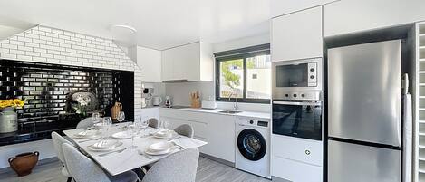 Una cocina moderna con armarios blancos, horno empotrado, refrigerador de acero inoxidable y comedor con mesa dispuesta para cuatro.