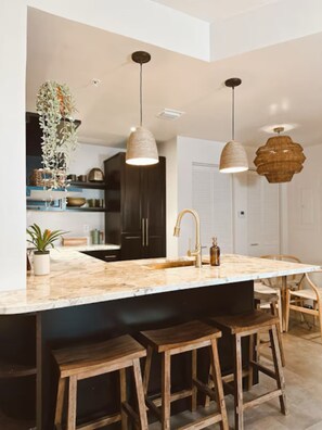 Une cuisine moderne avec un plan de travail en marbre, des tabourets en bois, des luminaires suspendus et une plante en pot.