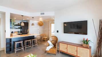 Un salon moderne doté d’un grand téléviseur à écran plat fixé au mur, d’un coin repas avec une table et des chaises, et d’une cuisine avec un comptoir-bar et des tabourets.