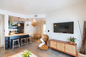 Un salon moderne doté d’un grand téléviseur à écran plat fixé au mur, d’un coin repas avec une table et des chaises, et d’une cuisine avec un comptoir-bar et des tabourets.