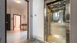 Un ascensore con interni metallici e pavimento piastrellato che conduce a una stanza con un tavolo di legno e una porta con scritto "Sole".