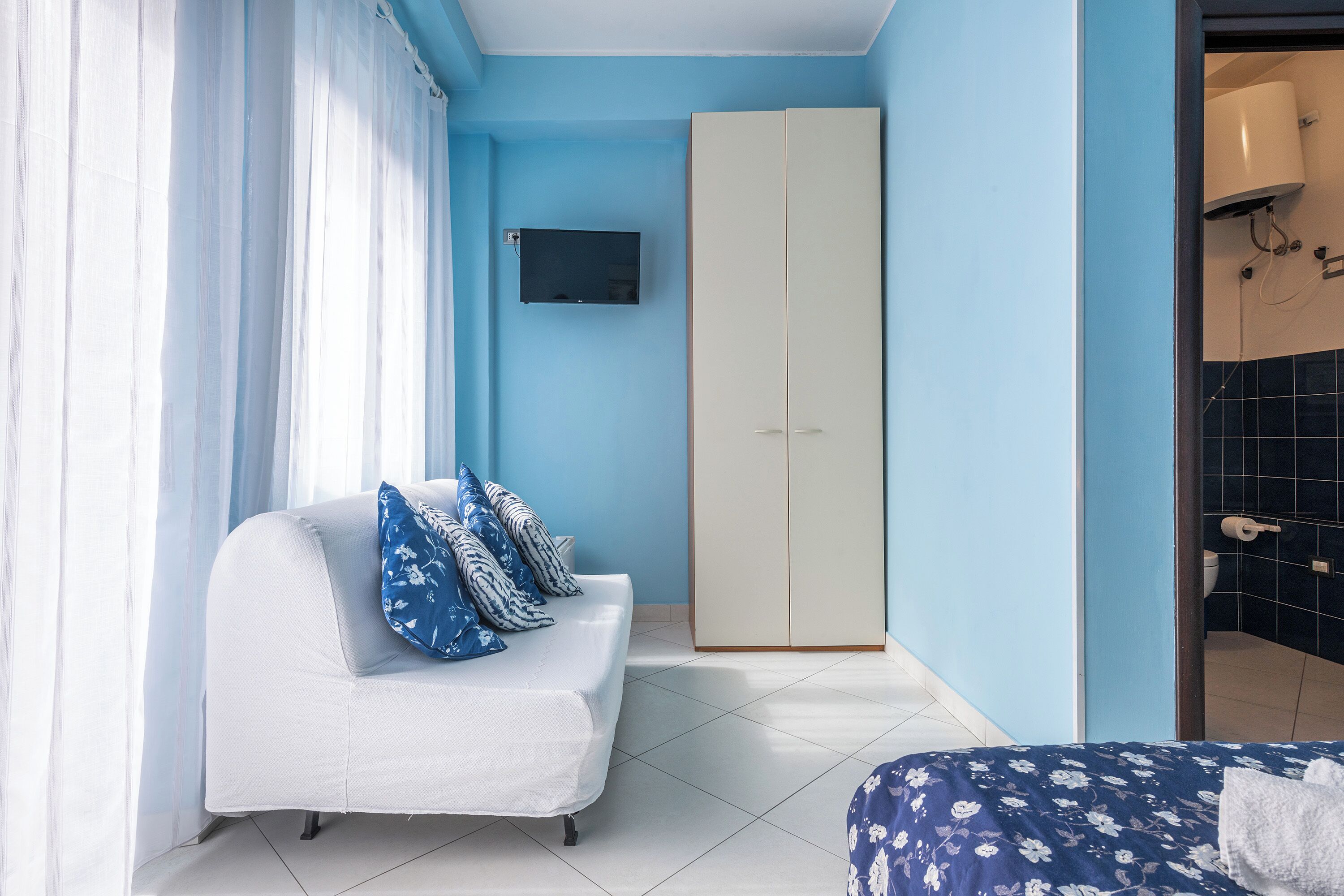 A bedroom with a white sofa, a bed with a floral bedspread, a wardrobe, and a television on the wall.
