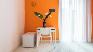 Una stanza con una parete arancione, una scrivania bianca, una sedia bianca, un piccolo elettrodomestico e un vaso con fiori.