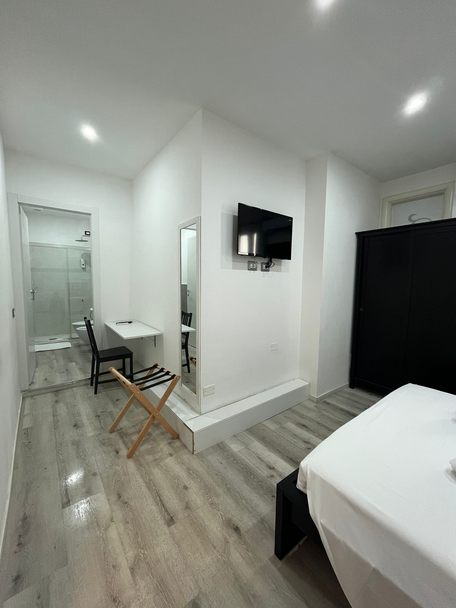 A modern hotel room with a bed, a desk, a chair, a TV mounted on the wall, and a wardrobe.