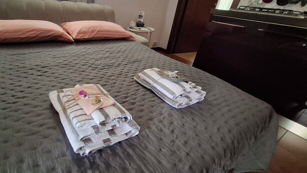 A neatly made bed with a grey quilt, two pink pillows, and two folded towels with a striped pattern and a small pink item on them. - Vacation House "al Vicoletto" (Pratola Peligna)