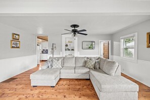 Un salon spacieux avec un grand canapé d’angle, un plancher en bois, des murs blancs et un ventilateur de plafond.