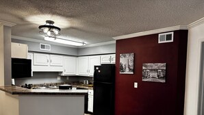 Une cuisine avec des armoires blanches, un réfrigérateur noir et un plan de travail en marbre. On y trouve deux tabourets et une poubelle.