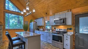 A cabin-style kitchen with a central island, stainless steel appliances, and a view of the forest through large windows. - Willow Falls (Blue Ridge)