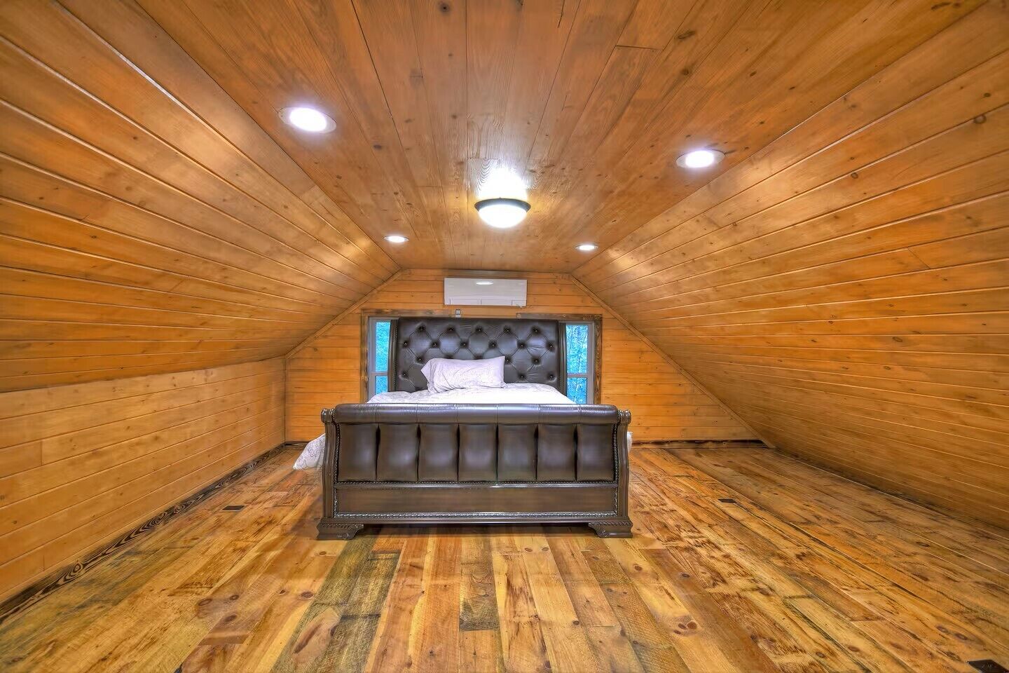 A bedroom with a wooden sloped ceiling, a bed with a tufted headboard, and a window on one wall.