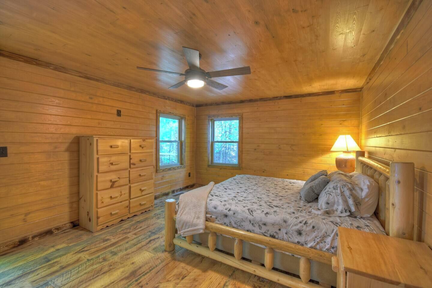 A wooden cabin bedroom with a bed, dresser, two windows, a ceiling fan, and a nightstand with a lamp.
