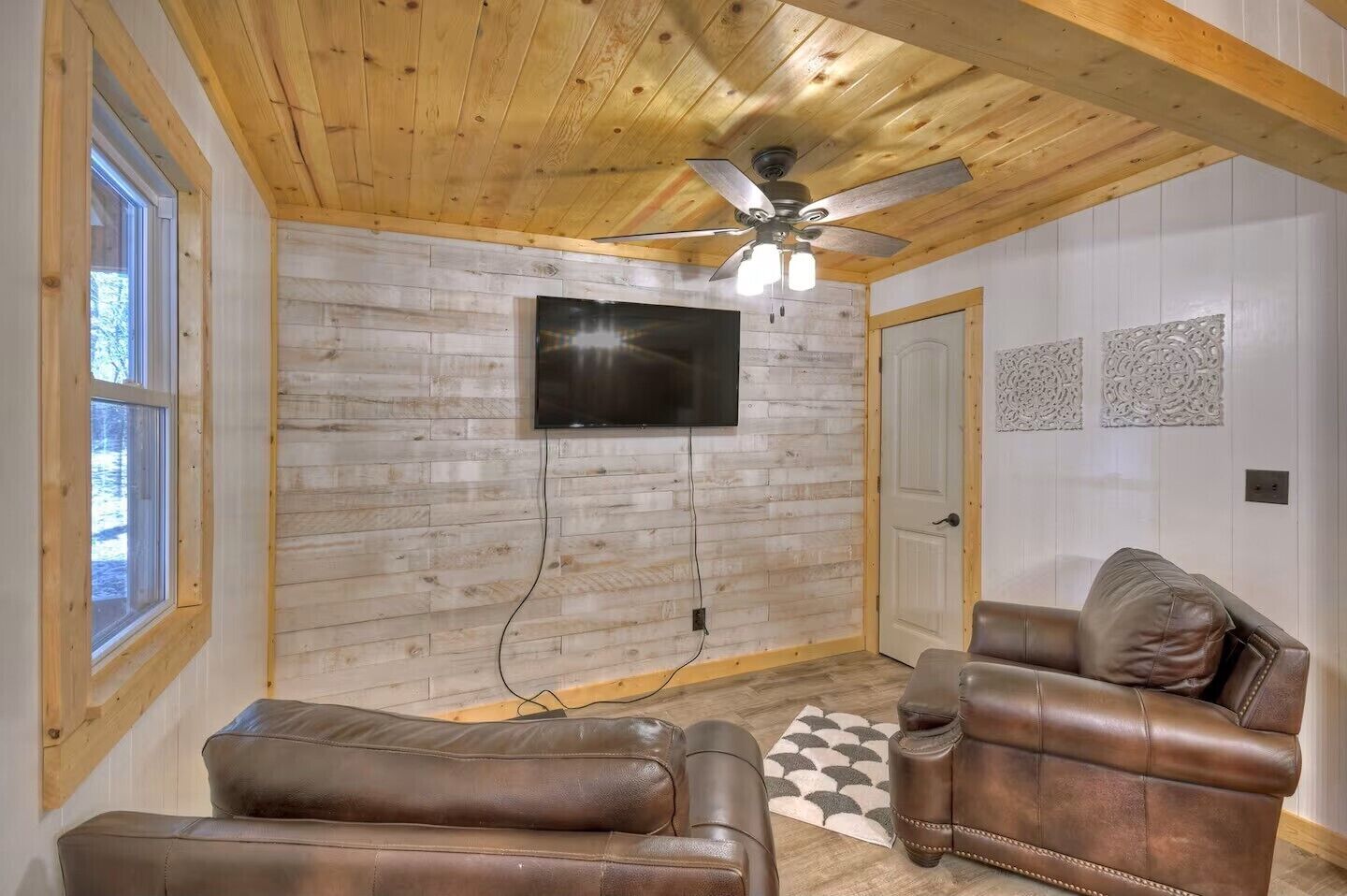 Une pièce avec un plafond en bois, un ventilateur de plafond, un téléviseur fixé au mur, une porte et deux canapés en cuir.