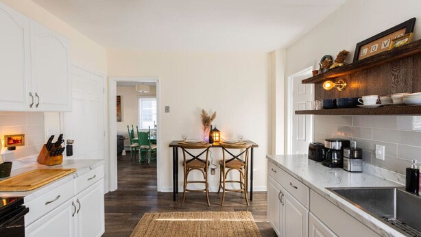 A modern kitchen with white cabinets, a wooden dining table with two chairs, a coffee machine, and a shelf with decorative items. - Private Modern 3 Bed Apt Close to Downtown- 1st Flr (Torrington)