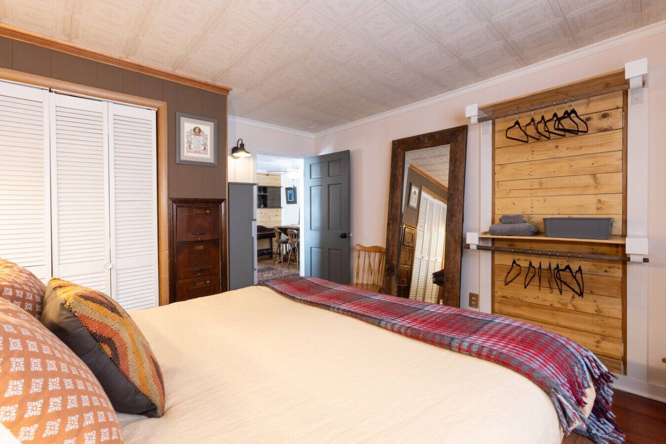 Quarto com cama, guarda-roupa, espelho e parede de madeira com roupas penduradas.