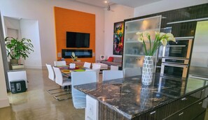 A modern kitchen with a marble countertop, stainless steel appliances, and a dining area with white chairs and a colorful table. - Elysian Haven - Modern Urban Oasis in a Peaceful Neighborhood (San Antonio)