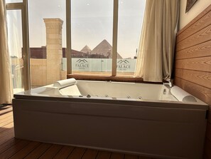 A modern bathroom with a large bathtub, a wooden wall, and a view of the pyramids through a window. - Ra Palace Pyramids View (Giza)