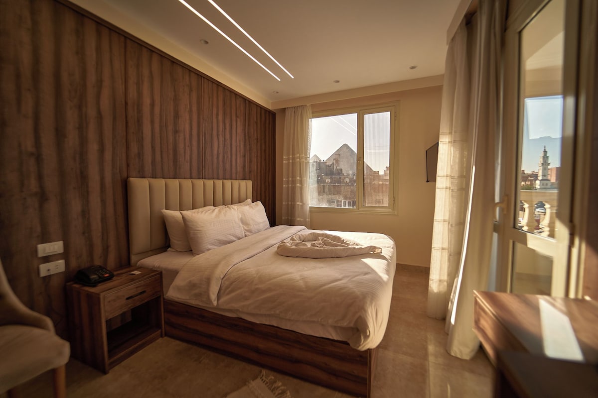A modern bedroom with a large bed, wooden paneling on one wall, a bedside table with a phone, and a window with a view of a building.