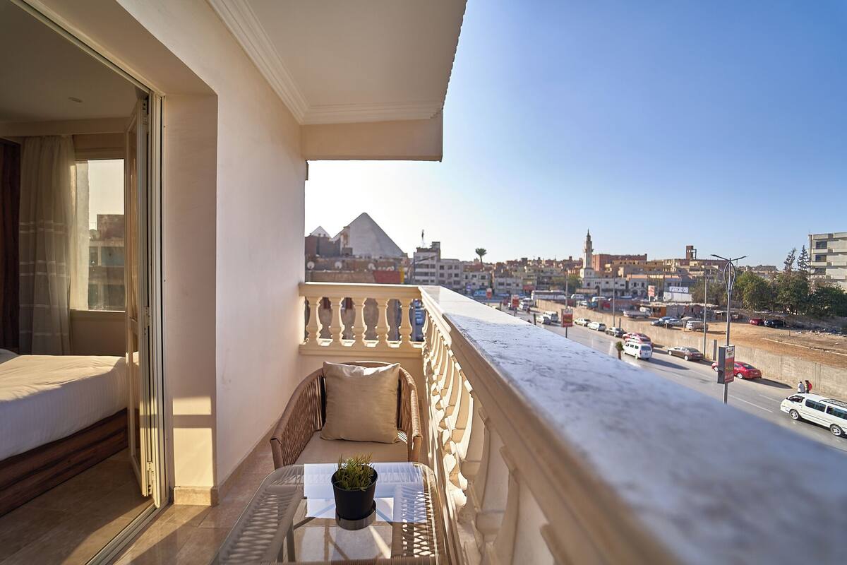 A balcony with a wicker sofa, a small table with a plant, and a view of a cityscape including a pyramid.
