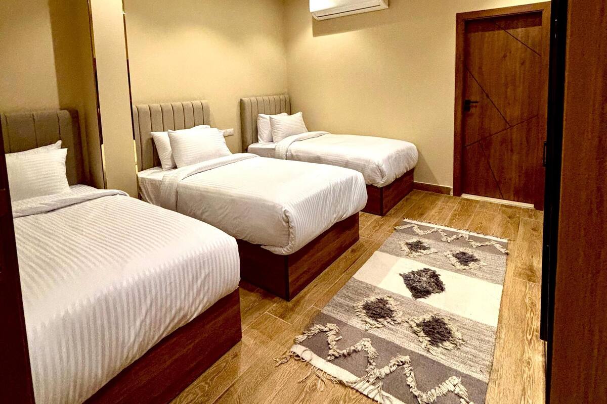 A hotel room with three beds, a wooden floor, a patterned rug, and an air conditioning unit on the wall.