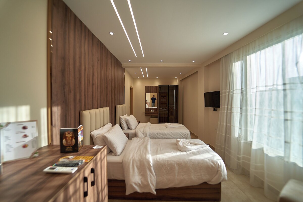 A hotel room with two beds, a wooden desk, a flat-screen TV, and a large window with sheer curtains.