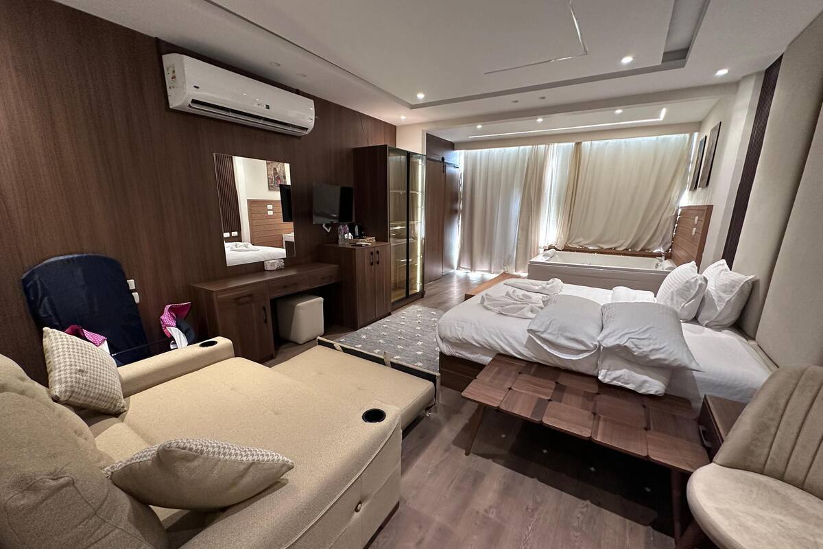 A hotel room with a sofa, a bed, a desk, a mirror, and an air conditioning unit.