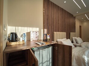 A modern hotel room with a wooden desk, a coffee maker, a water bottle, a menu, and a minibar.