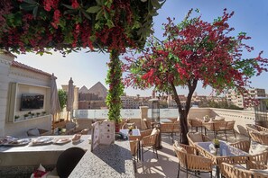 An outdoor seating area with tables and chairs, a menu stand, and a view of the pyramids in the background.