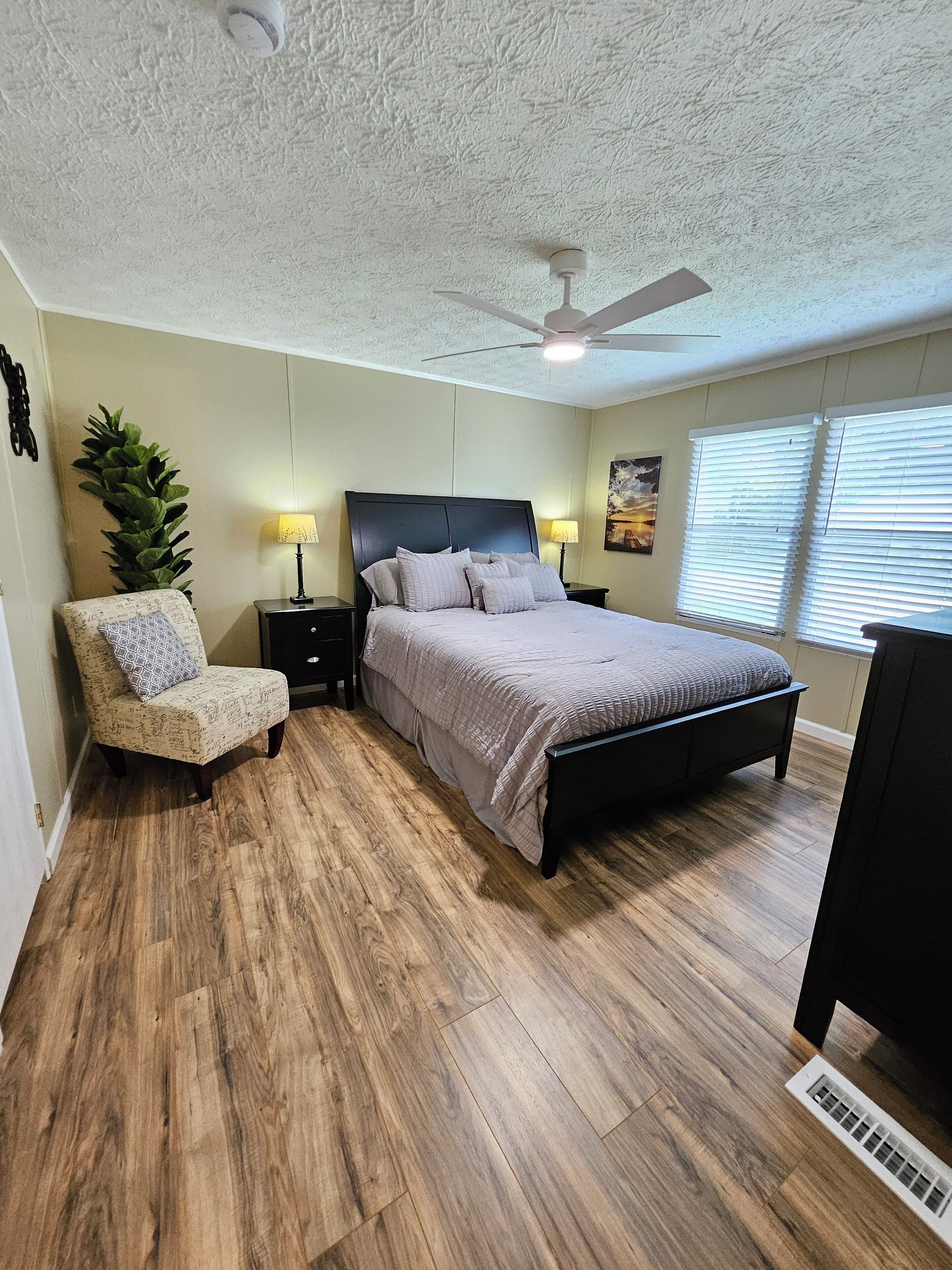 A bedroom with a bed, a chair, a nightstand, a ceiling fan, and a window with blinds.