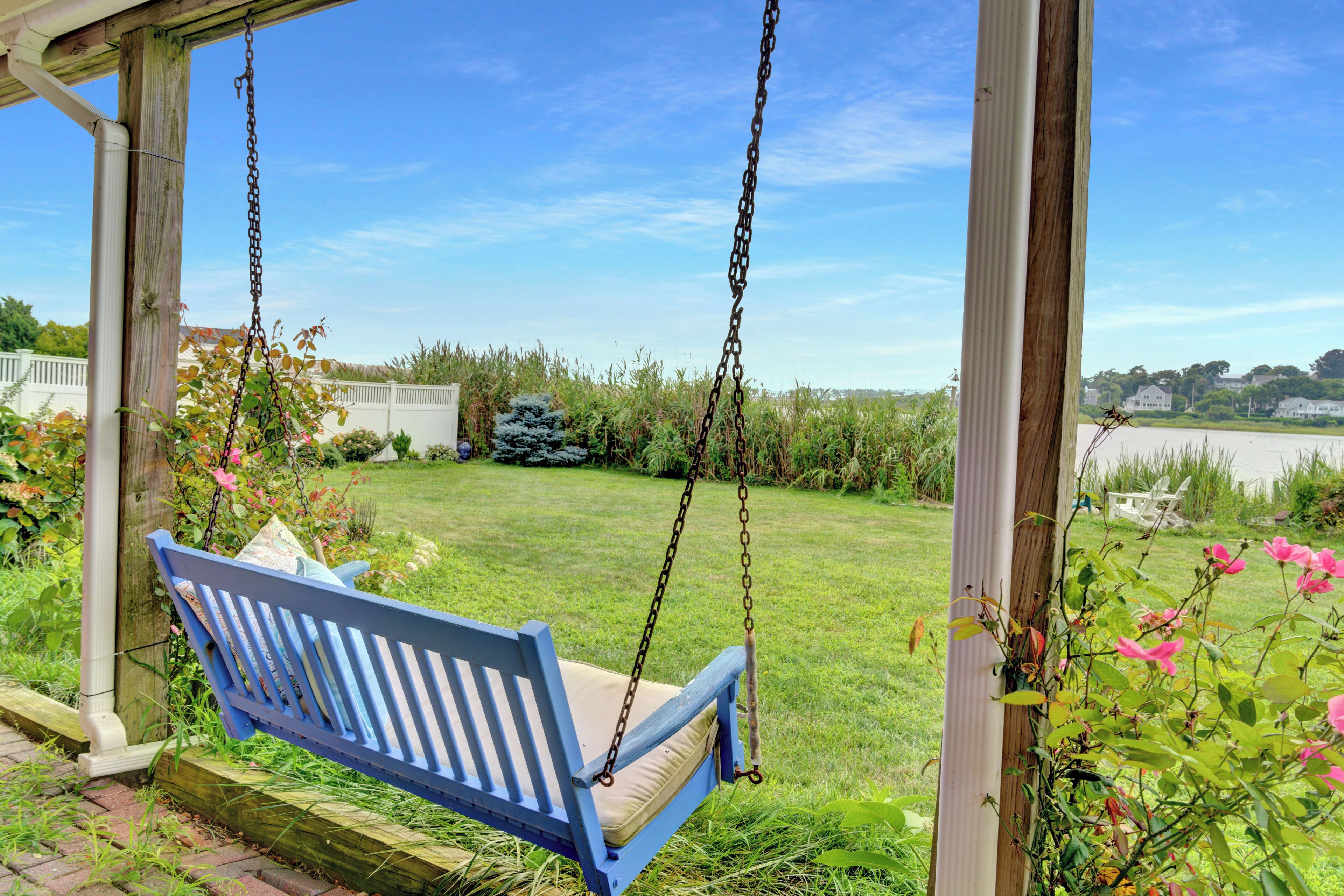 A blue wooden swing chair suspended by chains, set in a covered outdoor area with a grassy lawn and a view of a residential neighborhood.