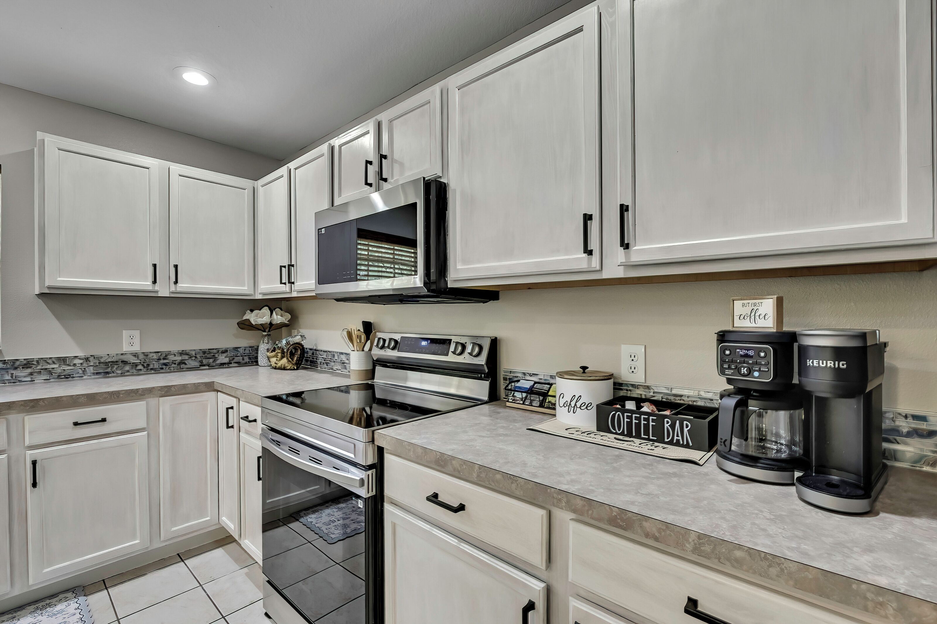 Una cocina moderna con armarios blancos, horno empotrado, microondas y una estación de café con máquina Keurig.